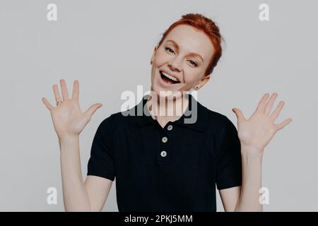Tolle Neuigkeiten. Studioaufnahme einer jungen, aufgeregten, rothaarigen Frau, die Handflächen hebt und laut ausruft, während sie isoliert auf grauem Hintergrund posiert und mut hält Stockfoto
