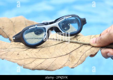 Schwimmbrille auf trockenem Blatt mit blauem Pool im Hintergrund, der das Konzept der Sommeraktivitäten zeigt Stockfoto