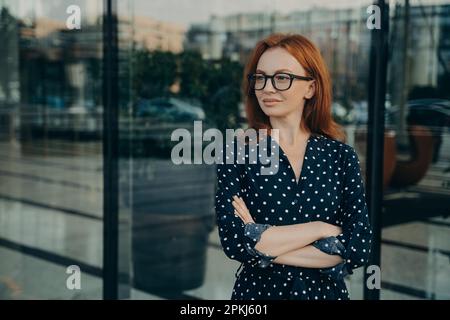 Die horizontale Aufnahme einer rothaarigen europäischen Frau schaut weg, hält die Arme zusammengeklappt und macht Pläne, um über zukünftige Karriere in schwarzer Polka d zu träumen Stockfoto