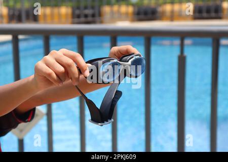 Schwimmbrille in der Hand mit einem Pool im Hintergrund, der das Konzept der Sommeraktivitäten im Freien zeigt Stockfoto