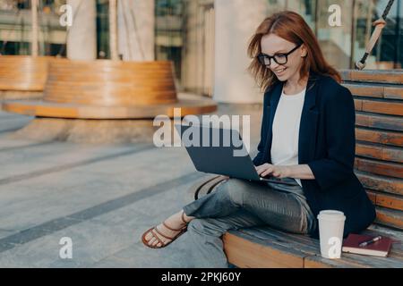 Fröhliche Freiberuflerin Tastaturen auf Laptop-Computer hat neue Ideen für die Arbeit trägt Brille formelle Kleidung sitzt auf einer Holzbank draußen mit Mitnahme Stockfoto