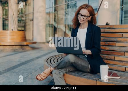 Die intelligente Rothaarige surft draußen auf dem Laptop und macht das Projekt online in eleganter Kleidung auf der Bank. Getränke zum Mitnehmen Kaffee genießt einen guten Tag. Stockfoto