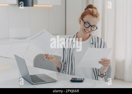 Lässig gekleidete, aufgeregte rothaarige Frau, die zu Hause am Tisch sitzt, sich glücklich fühlt, nachdem sie Inlandsrechnungen überprüft hat, Dokumente mit Banknoti in der Hand hat Stockfoto