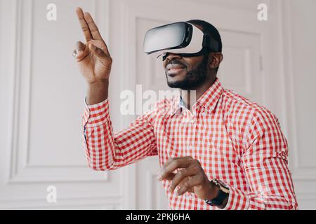 Ein begeisterter, lächelnder afrikanischer Mann in einer VR-Headset-Brille genießt die virtuelle Realität online und probiert die Cyber-World-Simulation bei der Arbeit aus Stockfoto