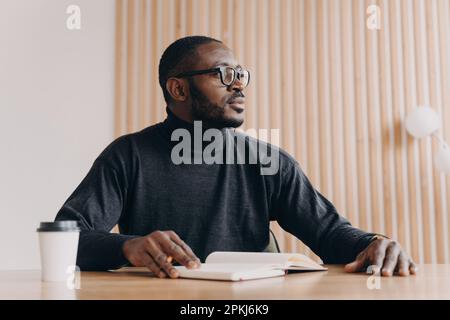 Junger, aufmerksamer afroamerikanischer Geschäftsmann, der mit Stift und Agenda am Schreibtisch sitzt und mit Nachsicht wegblickt. Milleniales birassisches Unternehmertum Stockfoto