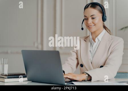 Glückliche Managerin des Callcenters, die in einem stilvollen Büro sitzt und Kunden berät, während sie an einem Laptop arbeitet, erfolgreiche Geschäftsfrau in mir Stockfoto