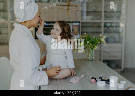 Ein hübsches, lockiges kleines Mädchen berührt die Nase ihrer Mutter, gekleidet in weißem, weichem Morgenmantel, sitzt auf dem Tisch mit Kosmetikprodukten und macht Make-up für mom Stockfoto