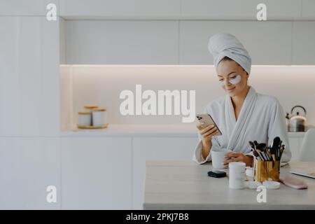 Glückliche junge europäische Frau im Bademantel Typen Textnachrichten im Online-Chat während der Kaffeepause wird Hautpflege Verfahren wendet Schönheitsbehandlung an Stockfoto