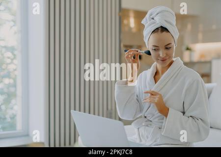 Foto einer jungen europäischen Frau trägt Gesichtspulver mit Kosmetikbürste auf, konzentriert in Laptop-Computer macht Videoanrufe und hat eigene Beauty-Blog-Posen in CO Stockfoto