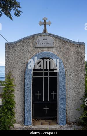 Saint Marina (Agia Marina) steht über der Tür. Blaue Kapelle mit Kreuz und Engel auf dem Dach, erbaut in einem wunderschönen kleinen Garten an der Küste. Verwandte Stockfoto