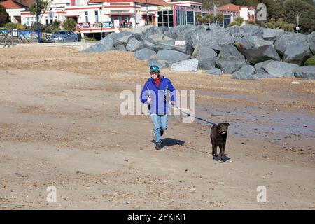 Eine Frau in Lila läuft mit einem Hund an einem Sandstrand Stockfoto