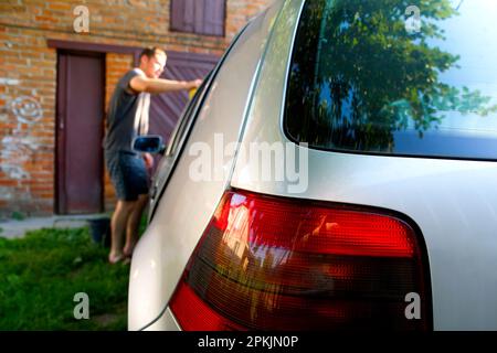 Autowaschanlage. Unschärfer-Mann wäscht graues Auto mit Schwammwäsche. Man hält die Mikrofaser in der Hand und poliert das Auto. Detailserie, Arbeiter putzt grau Stockfoto