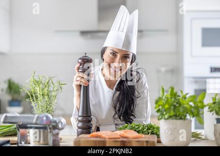 Die Chefköchin in Toque Blanche, weißem Hut und Uniform hält während der Zubereitung von Fisch- und Gemüsemahlzeiten eine Pfeffermühle. Stockfoto