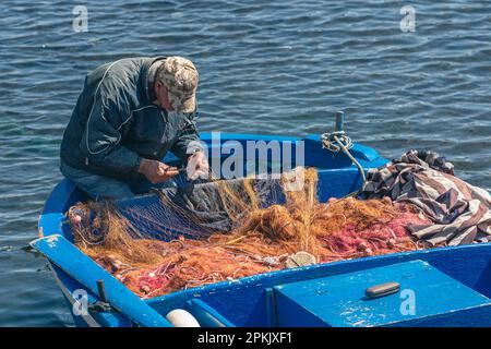 Ein Fischer mit einem Hut, der mit einer großen Nadel ein Fischernetz auf einem alten blauen hölzernen Fischerboot am Hafenkai in Bari, Apulien, Italien, repariert Stockfoto