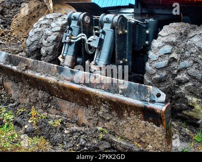 Radtraktor mit Schaufel räumt an bewölkten Regentagen den Boden frei. Schmutzige Bulldozer-Schaufel Nahaufnahme... Stockfoto