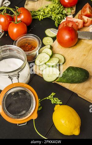 Reife geteilte Tomaten, Knoblauch, Gurkenscheiben, Paprika, eine Flasche Salz, ganze Zitronen, Olivenöl und ein Messer auf einem Tisch Stockfoto