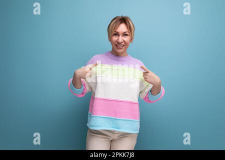 Die süße, bezaubernde blonde junge Frau in einem gestreiften Pullover zeigt fröhliche Emotionen des Glücks auf blauem Hintergrund Stockfoto