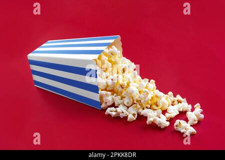 Leckeres Käsepopcorn, das aus einem blau gestreiften Eimer fällt, isoliert auf rotem Hintergrund. Streuung von Popcornkörnern. Filme, Kino und Enterta Stockfoto