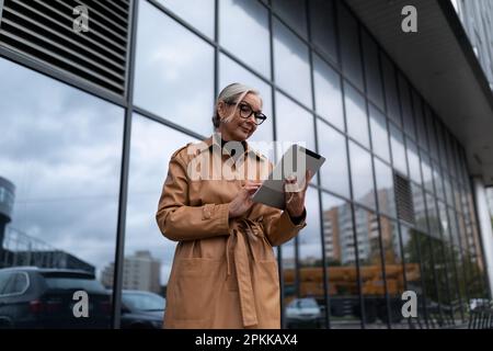 Selbstbewusste, reife Freiberuflerin mit grauen Haaren in Brillen, die mit einem Tablet in der Hand vor dem Hintergrund eines Business Centers steht Stockfoto