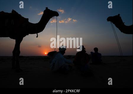 Thar Desert, Rajasthan, Indien-15.10.2019 : Silhouette von zwei Cameleers mit einer Touristin. Kamele in Sanddünen. Sonnenuntergang mit blauem Himmelshintergrund. Stockfoto