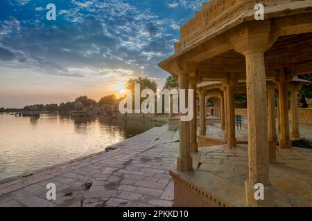 Chhatris und Schreine der hinduistischen Götter und Göttinnen am Gadisar-See, Jaisalmer, Rajasthan, Indien. Indo-islamische Architektur, Sonnenuntergang und bunte Wolken. Stockfoto