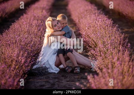 Ein Porträt von blonder Mutter mit kleinem Sohn sitzt im violetten Lavendelfeld. Junge Frau in weißem Kleid umarmt und küsst den Jungen liebevoll. Die Conce Stockfoto