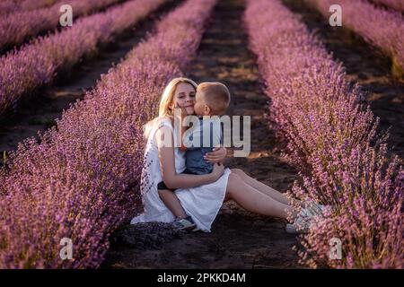 Ein Porträt von blonder Mutter mit kleinem Sohn sitzt im violetten Lavendelfeld. Junge Frau in weißem Kleid umarmt und küsst den Jungen liebevoll. Die Conce Stockfoto