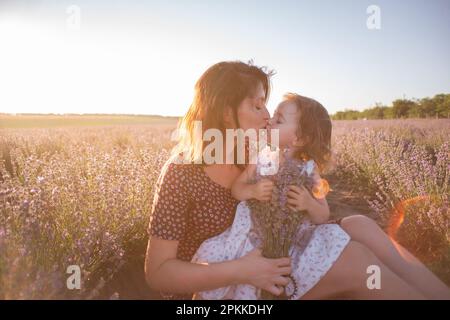 Porträt einer brünetten Mutter mit kleiner Tochter, die im violetten Lavendelfeld sitzt. Junge Frau in ländlichem Kleid umarmt sich liebevoll, küsst Mädchen. Die Konz Stockfoto