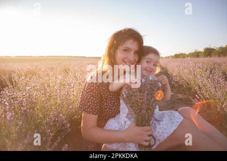 Porträt einer brünetten Mutter mit kleiner Tochter, die im violetten Lavendelfeld sitzt. Junge Frau in ländlichem Kleid umarmt sich liebevoll, küsst Mädchen. Die Konz Stockfoto