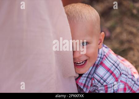 Nahaufnahme eines kleinen Jungen im karierten Hemd, umarmtes Mütterbein. Draufsicht auf ein lächelndes Kleinkind mit blauen Augen. Der Begriff des elterlichen Schutzes, t Stockfoto