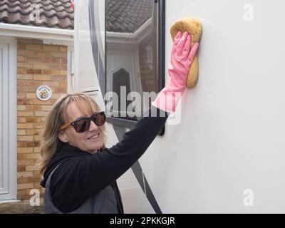 Lächelnd und in die Kamera eine attraktive Frau, die ein Wohnmobil besitzt, wäscht ihr Freizeitfahrzeug mit einem Schwamm. Sie trägt pinke Handschuhe und eine Sonnenbrille Stockfoto