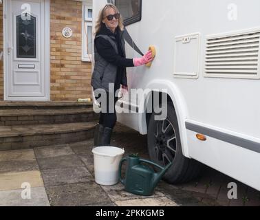 Eine Dame lächelt, während sie steht und ihr Wohnmobil mit einem Schwamm reinigt. Eimer und Gießkanne sind sichtbar. Stockfoto