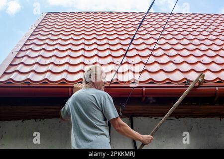 Ein reifer Mann reinigt die Rinne eines Abflussrohrs auf dem Dach seines Hauses von Herbstblättern und Schutt. Stockfoto