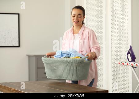 Junge Frau, die einen Korb voller sauberer Wäsche auf einen Holztisch im Haus legt Stockfoto