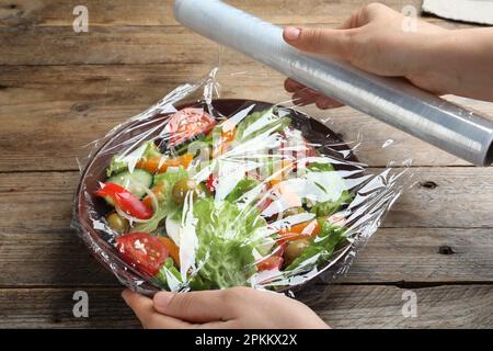 Frau, die Plastikfolie über eine Schüssel mit frischem Salat am Holztisch legt, Nahaufnahme Stockfoto