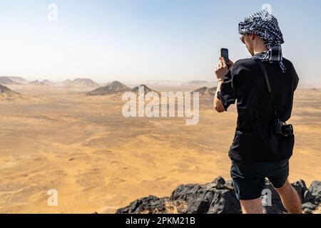 Ein Tourist, der ein Foto auf einem felsigen Felsvorsprung/Basalthügel in der Schwarzen Wüste in Ägypten macht Stockfoto