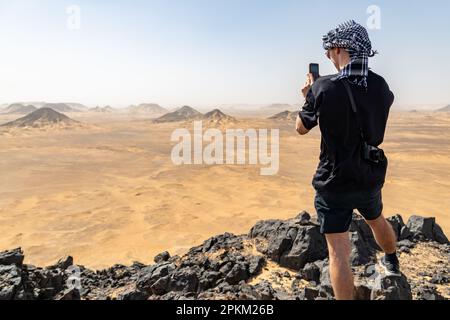 Ein Tourist, der ein Foto auf einem felsigen Felsvorsprung/Basalthügel in der Schwarzen Wüste in Ägypten macht Stockfoto