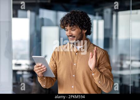 Erfolgreicher hispanischer Mann, der im Büro im Innenbereich am Arbeitsplatz steht, Mann, der aus der Ferne mit Kollegen per Videoanruf telefoniert, Geschäftsmann, der einen Tablet-Computer in der Hand hält, winkt mit einer Grußgeste. Stockfoto