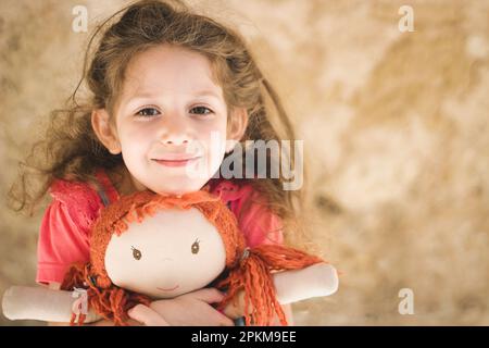 Ein niedliches, süßes, weißes Mädchen, das liebevoll eine geliebte Puppe mit Ingwerhaar in der Hand hält Stockfoto