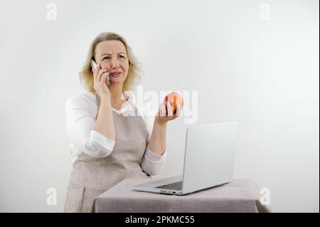 Eine Frau, die am Telefon spricht, ist verärgert und verärgert in ihren Händen. Sie hat eine richtige Ernährung mit einem roten Apfel vor dem Laptop Stockfoto