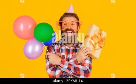 Glücklicher Mann mit Bart in lustigen Gläsern und Geburtstagsmütze mit Geschenkbox und Ballons. Ein hübscher Kerl feiert Geburtstag mit bunten Luftballons und Stockfoto