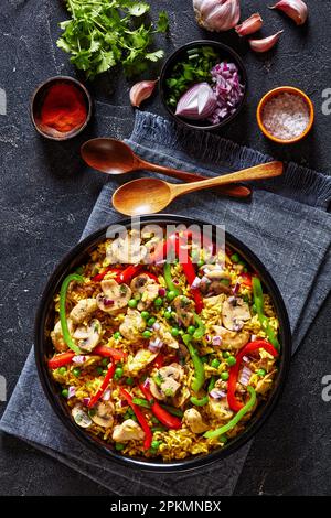 Hähnchenpilzpaella mit süßer Paprika, grünen Erbsen und Gewürzen in schwarzer Schüssel auf Betontisch mit Holzlöffeln und Gewürzen, vertikale Sicht von Stockfoto