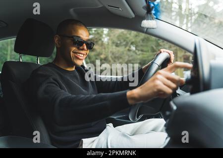 Fröhlicher junger Mann mit Sonnenbrille sitzt im Auto und macht die Musik an, mittlere Volltreffer. Hochwertiges Foto Stockfoto