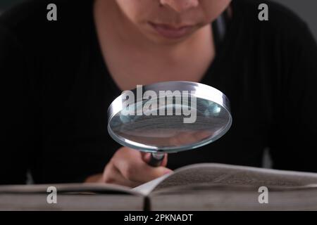Nahaufnahme einer Frau, die durch eine Lupe auf ein Lehrbuch schaut. Vergrößerungsglas in der Hand und offenes Buch auf dem Tisch. Bildungs- und Forschungskonzept. Stockfoto