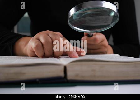 Nahaufnahme einer Frau, die durch eine Lupe auf ein Lehrbuch schaut. Vergrößerungsglas in der Hand und offenes Buch auf dem Tisch. Bildungs- und Forschungskonzept. Stockfoto