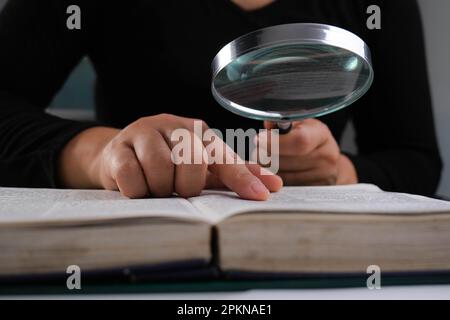 Nahaufnahme einer Frau, die durch eine Lupe auf ein Lehrbuch schaut. Vergrößerungsglas in der Hand und offenes Buch auf dem Tisch. Bildungs- und Forschungskonzept. Stockfoto