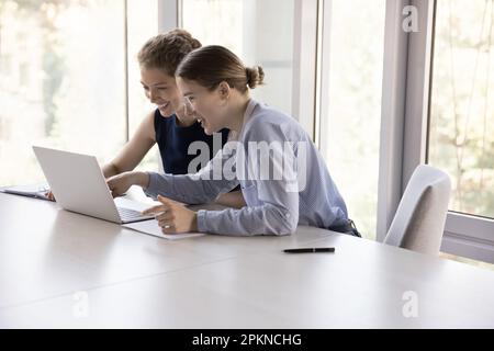 Zwei fröhliche junge Angestellte, die ihren Arbeitsplatz teilen und eine Online-Anwendung nutzen Stockfoto