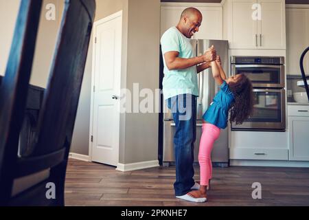 Diese Familie weiß, wie man Spaß hat. Ein glückliches kleines Mädchen, das auf den Füßen ihres Vaters balanciert, während sie zusammen in der Küche herumlaufen. Stockfoto