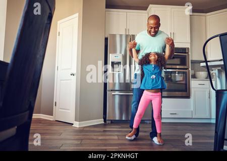 Gemeinsam Fortschritte machen. Ein glückliches kleines Mädchen, das auf den Füßen ihres Vaters balanciert, während sie zusammen in der Küche herumlaufen. Stockfoto