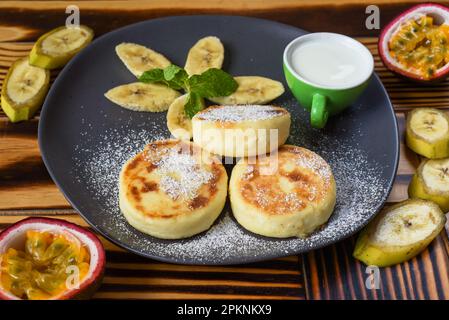 Syrniki- oder Käsepfannkuchen mit Sahne Sauce und Bananen auf einem Teller auf einem Holztisch aus nächster Nähe Stockfoto